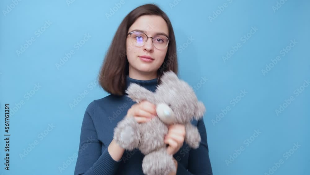 Video portrait of a cheerful student girl hugging, twisting a gray bear toy on a blue background, 4K. Favorite toy. Antistress for adults Video portrait of a cheerful student girl hugging, twisting a gray bear toy on a blue background, 4K. Favorite toy. Antistress for adults