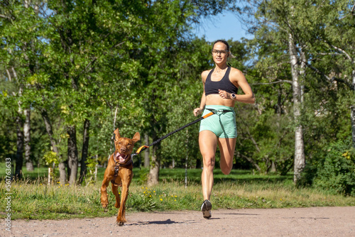 Sporty happy woman running with dog outdoor in public park summer. Canicross competition. Healthy life with active pet photo