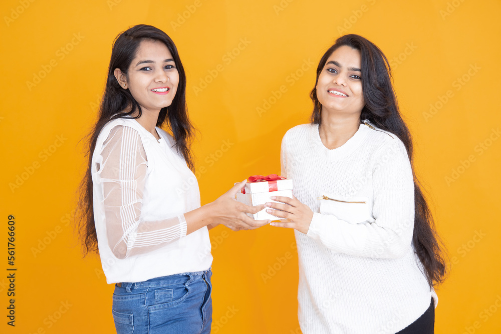 Happy young indian girls wearing white T-shirt exchange gift isolated over yellow orange background. studio shot. Festive celebration. Two happy brunette female friends exchanging presents, Happy young indian girls wearing white T-shirt exchange gift isolated over yellow orange background. studio shot. Festive celebration. Two happy brunette female friends exchanging presents,