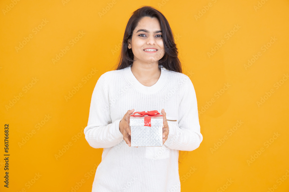 Happy young indian girl wearing white T-shirt holding gift looking at camera isolated over yellow orange background. studio shot. Festive celebration.