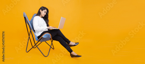 Young indian girl wearing white T-shirt using laptop while sitting on chair isolated over orange yellow background. Studio Shot, Copy space, Asian woman using computer.Technology concept.