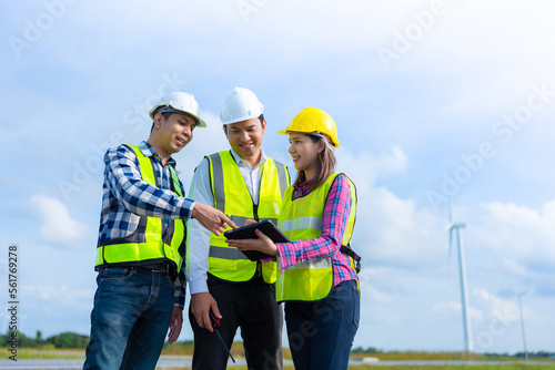 Team of engineers or architects, 3 men and women standing in a team meeting using a tablet Wear a helmet and a vest. with radio communication Wind turbines for electric power of industrial plants.