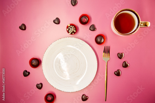 White mock up vintage plate with fork and cup of tea on pink gradient background. Chocolate hearts and decorated sweets. Romantic dinner, Valentines Day concept. View from above