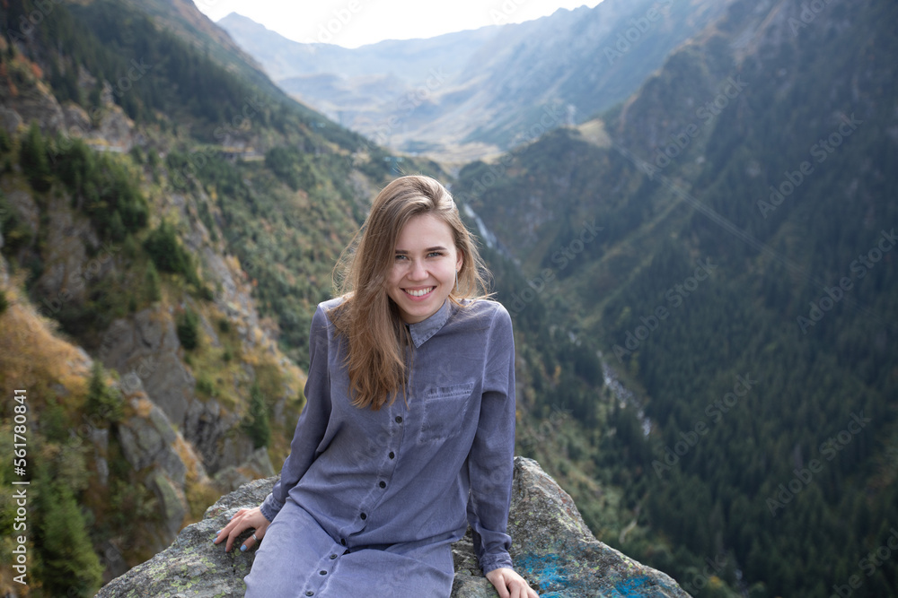 a girl with flowing hair and a beautiful smile is sitting on a stone in the mountains. a woman looks at the camera on the background of mountains