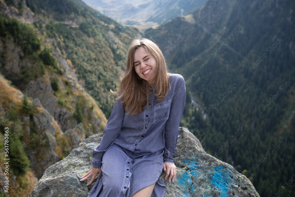 beautiful girl sits on a stone on the background of mountains in summer. closed her eyes and squints from the bright sun. rest in the mountains. hipster woman in dress