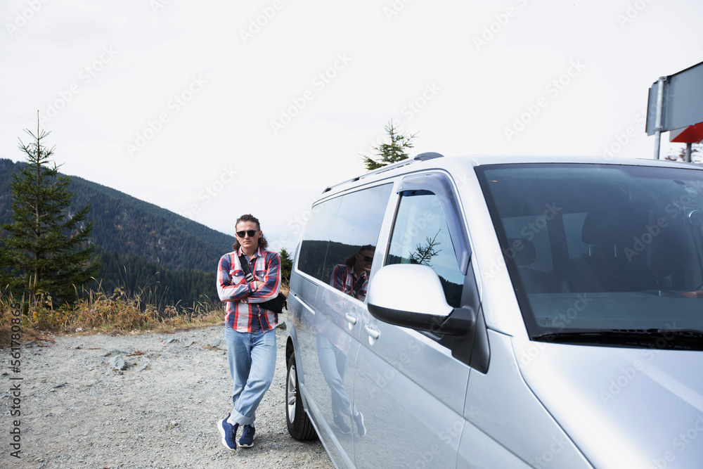 a hipster guy in sunglasses with his arms crossed in a checkered shirt is standing next to a gray minivan on a background of mountains. cool man with long hair. sunny day. general plan