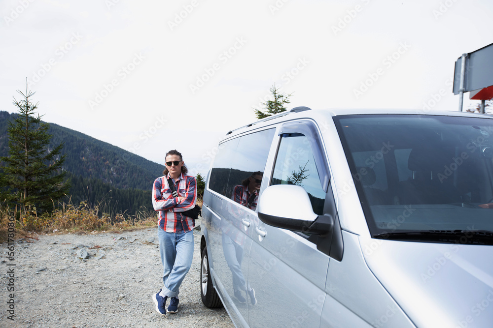 a hipster guy in sunglasses with his arms crossed in a checkered shirt is standing next to a gray minivan on a background of mountains. cool man with long hair. sunny day. general plan a hipster guy in sunglasses with his arms crossed in a checkered shirt is standing next to a gray minivan on a background of mountains. cool man with long hair. sunny day. general plan