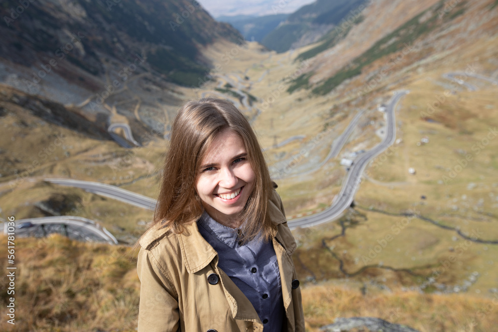 a beautiful girl in a light raincoat smiles and looks at the camera on the background of a serpentine in the mountains. Transfagarash highway. sunny day. Romania