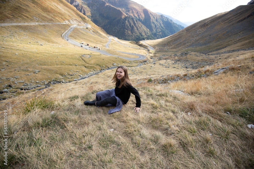 the girl laughs and sits on the dry grass in the big mountains against the background of the Sermantine road with cars. sunny day. Transfagarash mountains in Romania