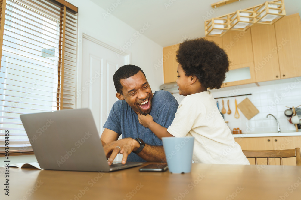 African American father working from home on laptop while his Cheerful son distracts him in kitchen. African American father working from home on laptop while his Cheerful son distracts him in kitchen.