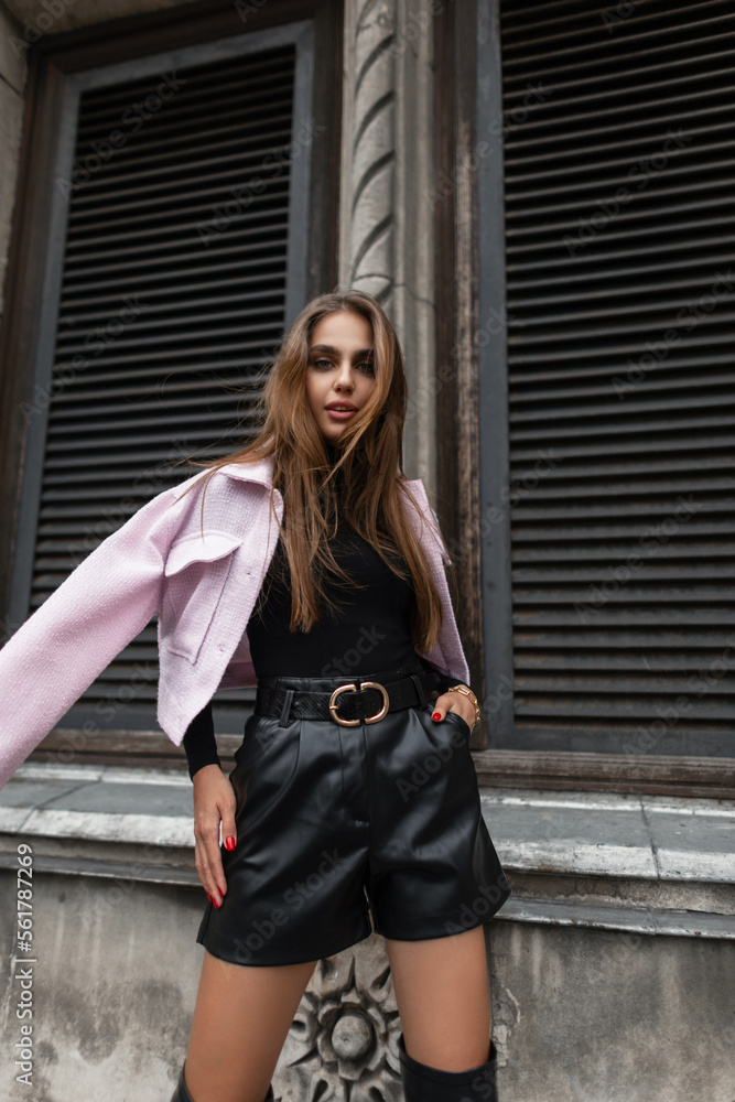 Beautiful stylish urban girl model in fashionable black outfit with a long sleeve shirt, leather shorts and a pink jacket on shoulders walks near a vintage building