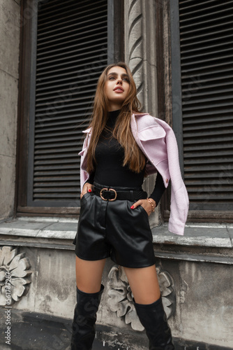 Stylish beautiful fashionable woman model in elegant black urban clothes with leather shorts and a sweater with a pink jacket on shoulders stands and poses near a vintage building with stucco