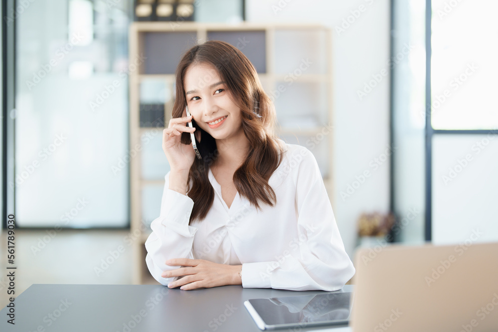 Portrait of a young Asian woman showing a smiling face as she uses his phone, computer and financial documents on her desk in the early morning hours Portrait of a young Asian woman showing a smiling face as she uses his phone, computer and financial documents on her desk in the early morning hours