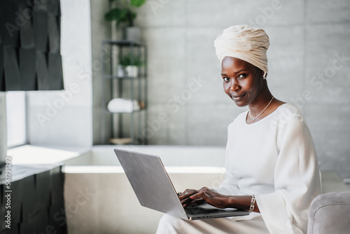 Photo Purposeful African woman in white turban works at home using laptop looks at camera