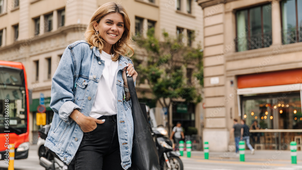 Beautiful woman smiling and looking at camera staying on the street. Fair-skinned blonde keep hand on pocket wearing white t-shirt and blue jacket. Streetstyle, lifestyle concept