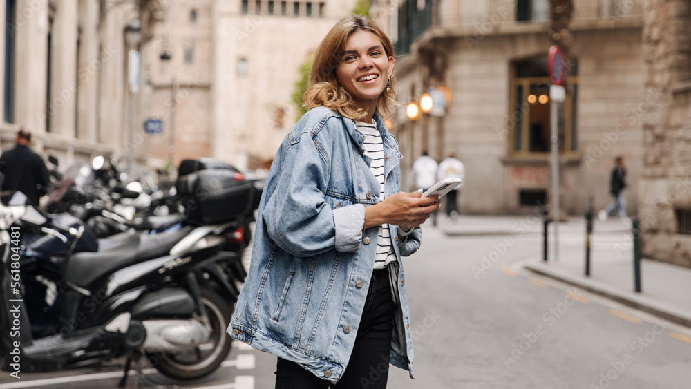 Naklejka premium Image of pretty teenager woman chatting on her smartphone and looking at camera. European smiling holding phone staying in the street. Concept of lifestyle, use technology 