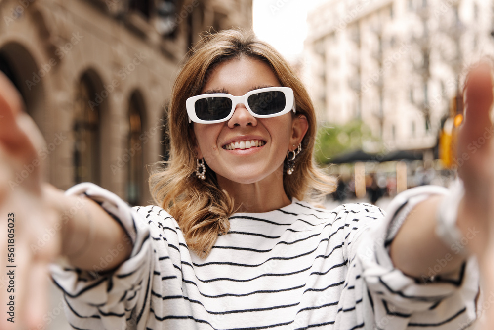 Cheerful european stylish woman reach hands to camera on the blur background. Fair-skinned blonde in sunglasses smiling. Emotional concept