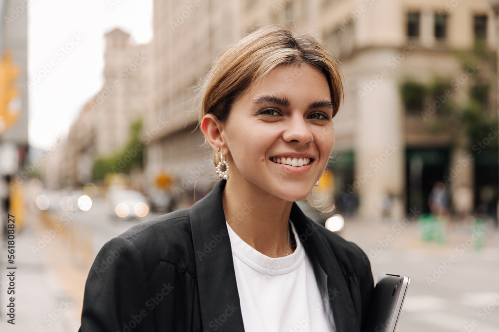 Portrait of happy charming woman look and smiling at camera on the street. Focused on blonde wearing black jacket, white t-shirt. Street style concept Portrait of happy charming woman look and smiling at camera on the street. Focused on blonde wearing black jacket, white t-shirt. Street style concept