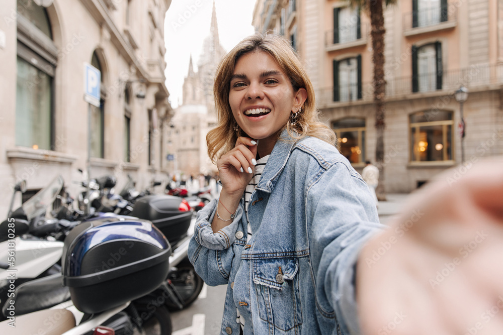 Fototapeta premium View of european happy woman keep hand on chin staying at the street. Smiling blonde looking and reach hand to camera. Different emotions concept