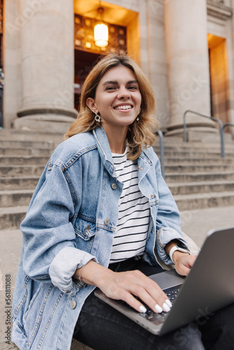 Portrait of attractive young woman sitting with laptop and looking at camera. Smiling european blonde in casual clothes. Concept of outdoots work time photo