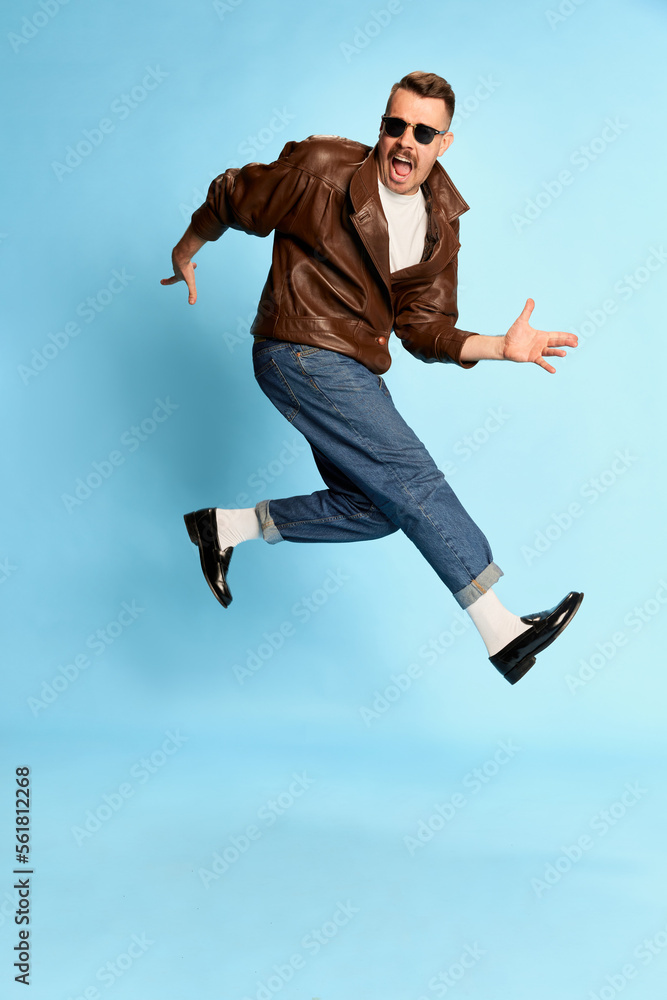 Fototapeta premium Portrait of brutal, handsome man in jeans, leather jacket and sunglasses posing, jumping over blue studio background. Concept of emotions, lifestyle