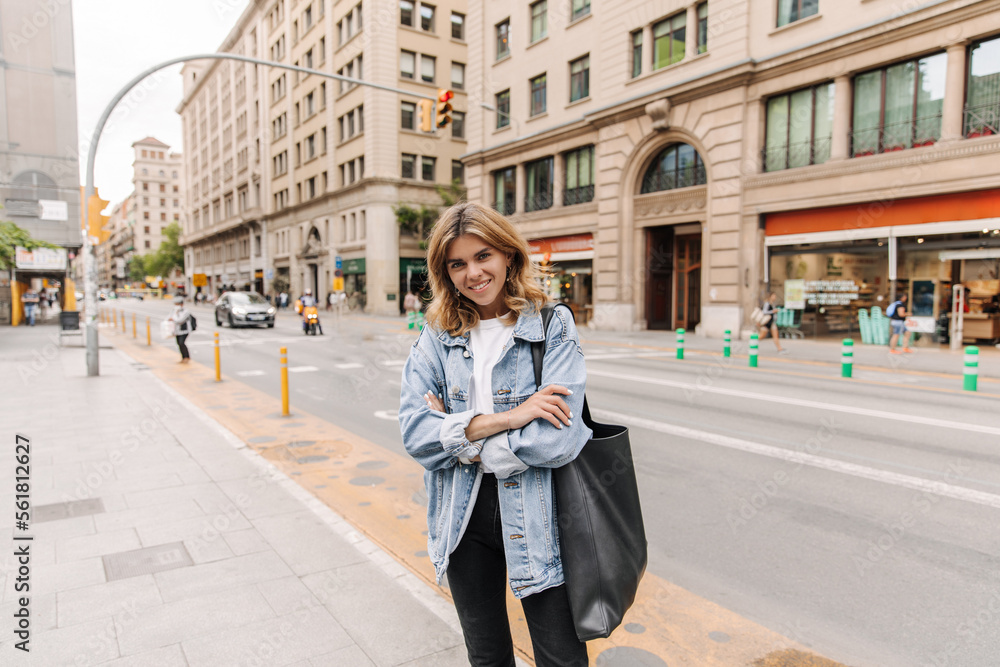 View of pretty stylish woman on the street smile at camera . Fair-haired caucasian lady keep crossed her hands staying with black bag. City style concept 