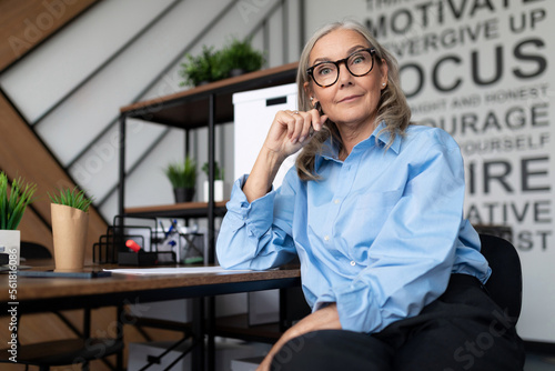 a mature businesswoman with a blue strict shirt sits against the backdrop of an office in glasses looking attentively into the camera