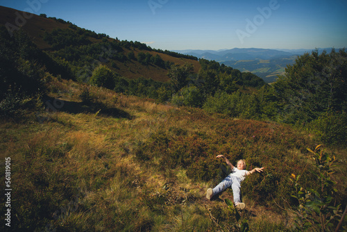 A little girl in blue pants and a light T-shirt lies in the grass in the mountains, in a hot summer. Around the grass, trees and blue sky with clouds.
