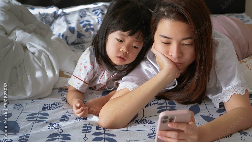 a little girl with her mom together watching over the screen of a smartphone while lying in prone position on the bed a little girl with her mom together watching over the screen of a smartphone while lying in prone position on the bed