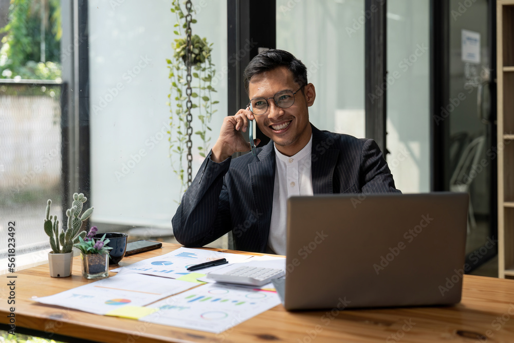 Businessman sitting at office desk in front of laptop hold mobile phone make pleasant business or informal call. Successful businessman looking at financial statistic shown on documents Businessman sitting at office desk in front of laptop hold mobile phone make pleasant business or informal call. Successful businessman looking at financial statistic shown on documents