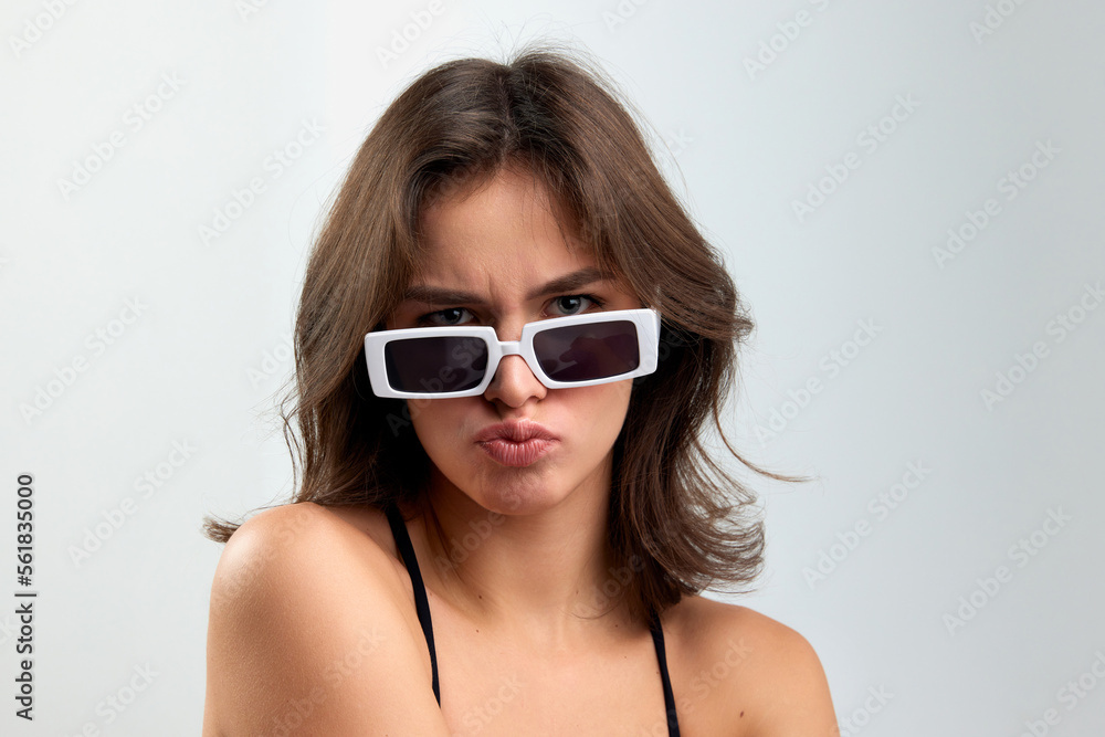 Doubts. Portrait of young stylish beautiful girl in fashionable eyewear isolated over grey background. Concept of beauty, skin care, cosmetology, fashion, makeup Doubts. Portrait of young stylish beautiful girl in fashionable eyewear isolated over grey background. Concept of beauty, skin care, cosmetology, fashion, makeup