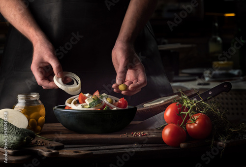 Cooking process of Greek Salad for a restaurant menu. Food composition of a Mediterranean cuisine in rustic style. Chef put the onion and olives in a black bowl and mixed of all vegetables in a salad.