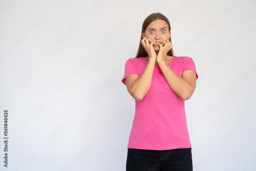 Scared young woman biting nails. Frustrated Caucasian female model with brown hair in pink T-shirt looking at camera with wide eyes, nervous about future. Anxiety concept Scared young woman biting nails. Frustrated Caucasian female model with brown hair in pink T-shirt looking at camera with wide eyes, nervous about future. Anxiety concept