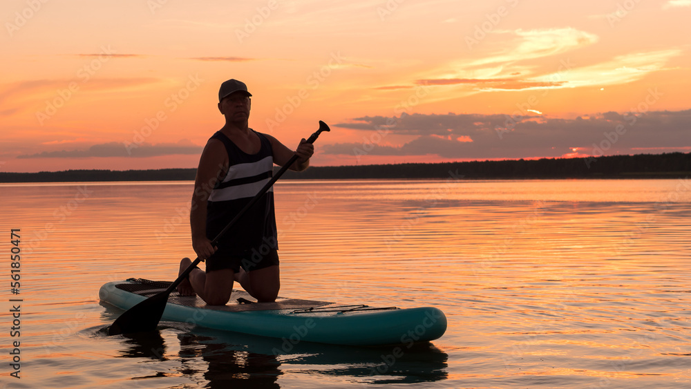 A man on a SUP board on his knees with an oar at sunset against a pink sky floats in the water of the lake.
