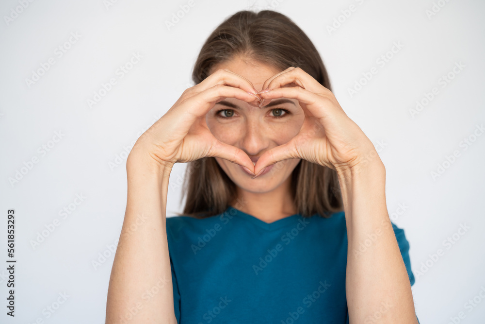 Happy Caucasian woman showing heart gesture. Portrait of pleased young female model with brown hair in blue T-shirt looking at camera, smiling behind heart shape. Love, romance concept Happy Caucasian woman showing heart gesture. Portrait of pleased young female model with brown hair in blue T-shirt looking at camera, smiling behind heart shape. Love, romance concept