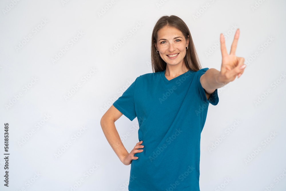Happy Caucasian woman showing victory sign. Portrait of cheerful young female model with brown hair in blue T-shirt looking at camera, smiling and greeting someone. Success, optimism concept