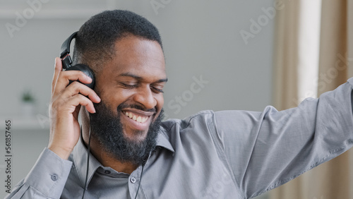 Happy African American man office worker ethnic businessman manager entrepreneur executive in headphones listening to music sitting at workplace funny relaxed guy enjoy favorite audio melody dancing