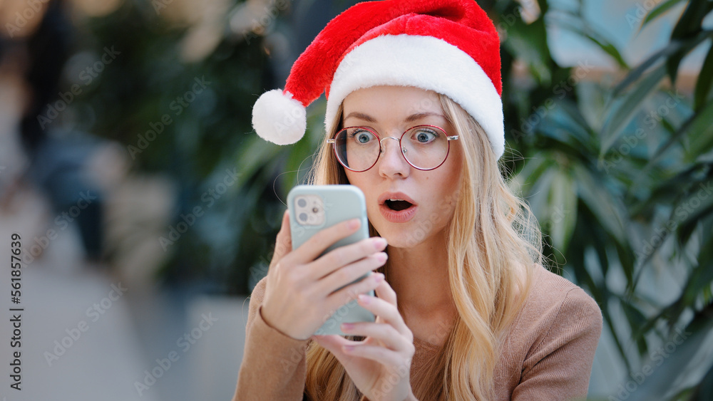 Surprised wonder astonish shocked amazed Caucasian woman girl lady in Santa Claus Christmas hat and eyeglass looking at phone mobile win victory offer achievement New Year celebration winning discount