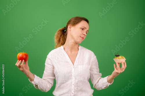 A middle-aged woman with an apple and a cake is tormented by her choice. On a green background. The concept of choosing the right healthy food.