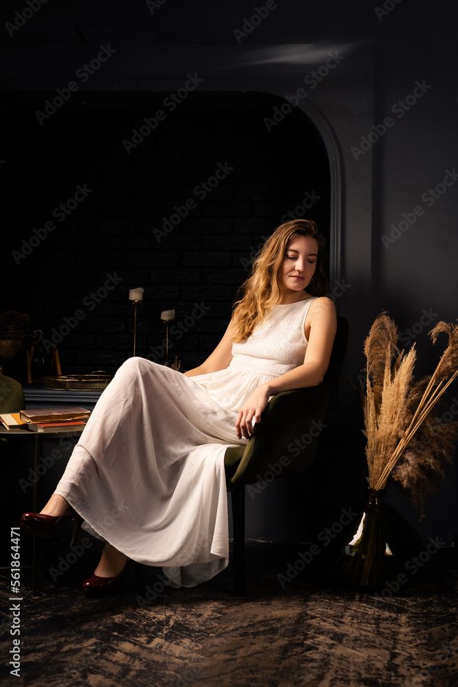 A young woman in a white dress sits thoughtfully in an armchair in a dark room. Solitude, introspection concept.