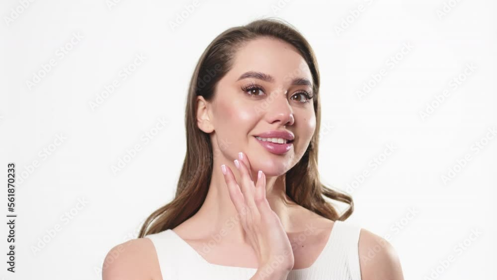 A slender young beautiful caucasian woman with dark hair in a white top turns to the left, touching her jaw line and smiling broadly at the camera on a white background. Moisturizer ad