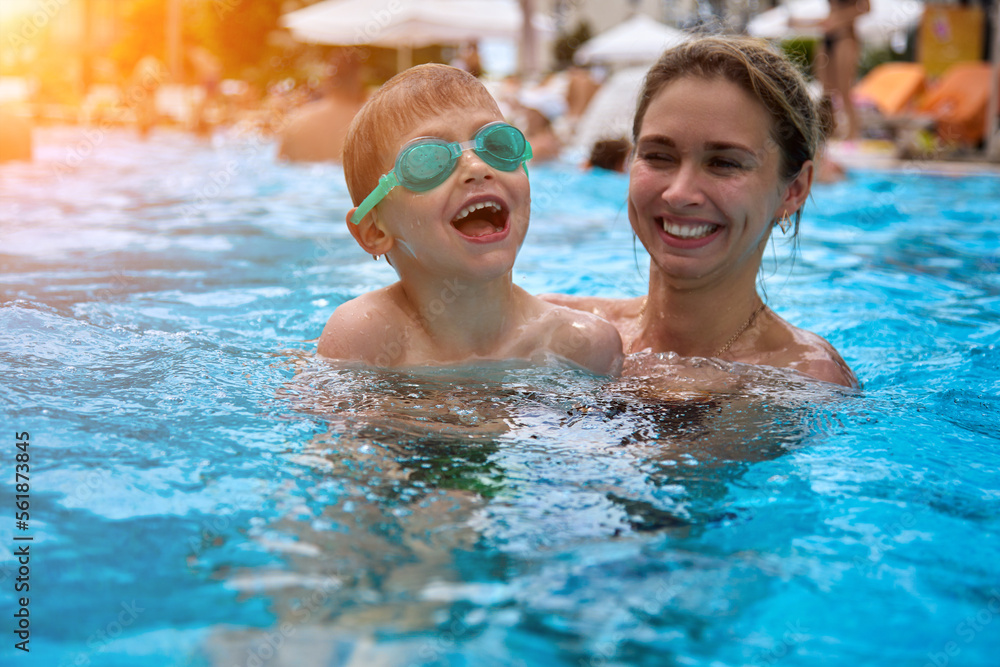 Cute young mother teaches her beloved son to swim in the pool in bathing glasses at the resort in the summer. Woman smiles, swims with her son in an outdoor pool on a summer vacation