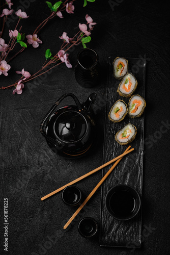 Baked sushi roll, chopsticks, sake bottle, cups and black teapot on black background 