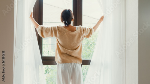 Happy Asian girl with white cream pajamas open white curtain on window looking outside feel fresh with peaceful morning and fresh air on weekend in bedroom at home. Female morning lifestyle concept.