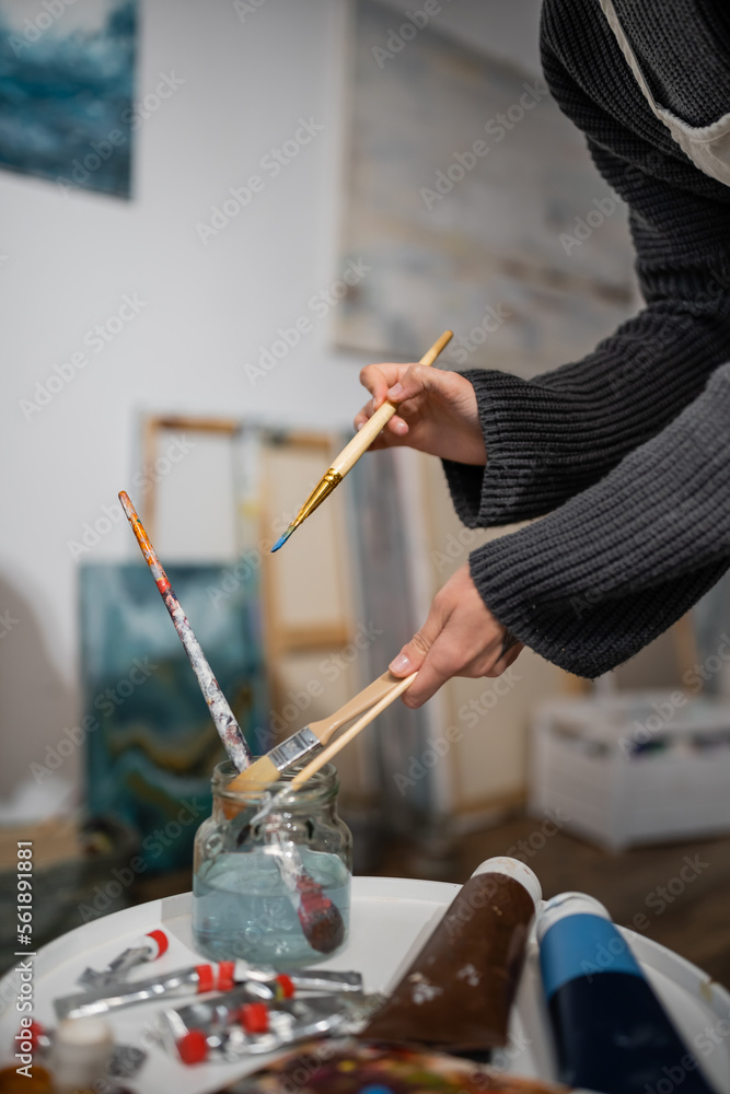 Fototapeta premium Cropped view of artist taking paintbrushes from jar with water in workshop