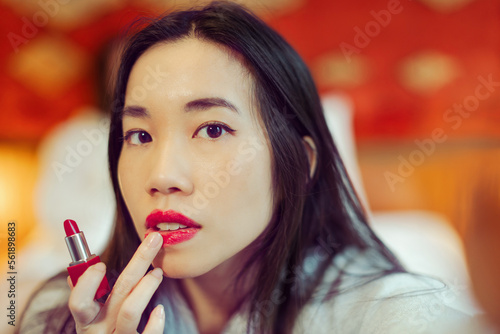 attractive south east asiatic woman in bathrobe looking in hotel room mirror putting red lipstick on her lips - close-up view