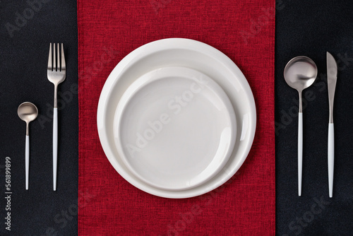 Festive place setting with red napkin. Empty plates and silver cutlery on dark black background. Top view. Dining table in restaurant. Card or menu template, flat design. Tableware, clean crockery.