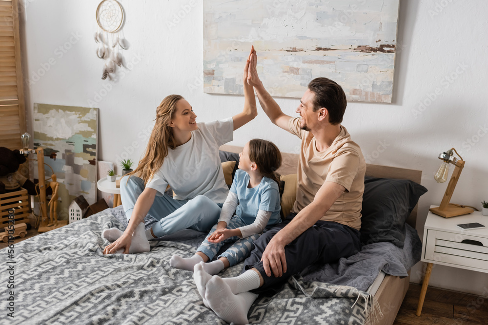 © LIGHTFIELD STUDIOS - cheerful kid looking at happy happy parents giving high five while sitting on bed.