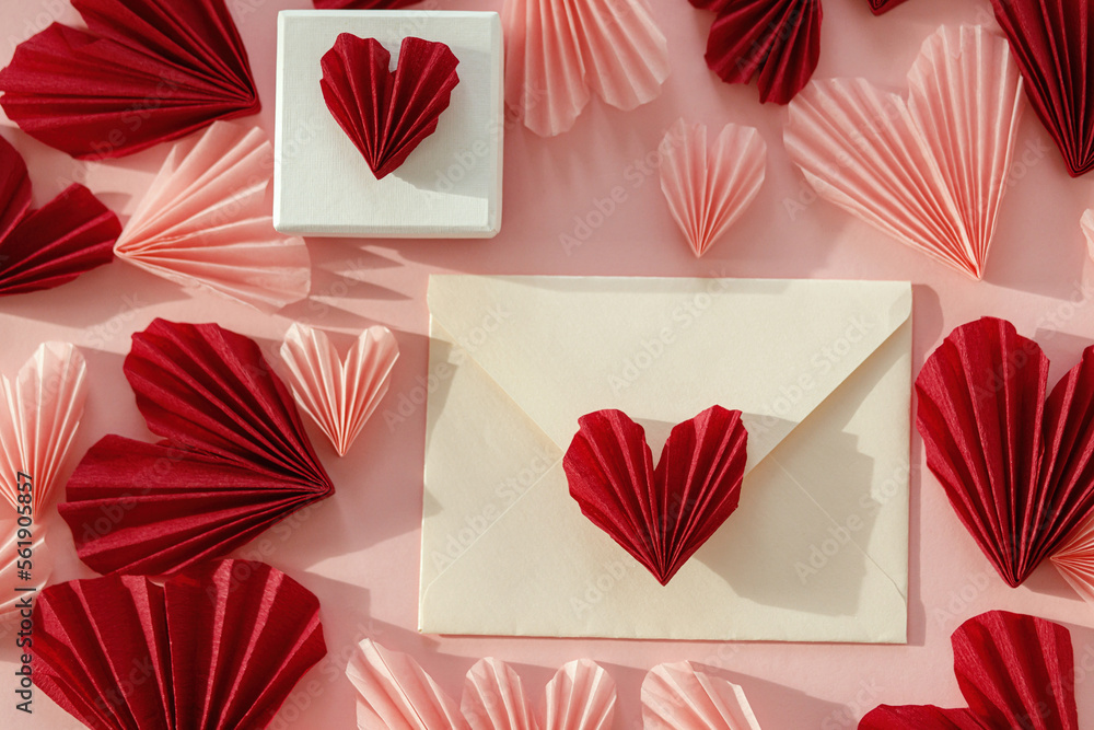 Obraz premium Happy Valentine's day! Stylish envelope with pink and red hearts and gift flat lay on pink paper background. Modern valentine card with heart cutouts. Love letter. Creative composition