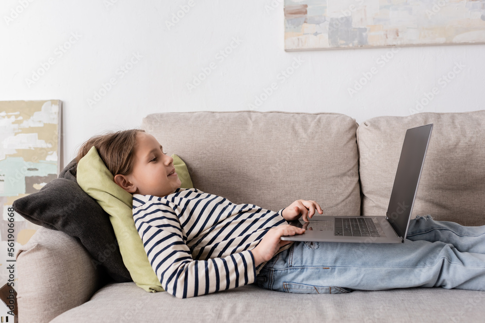 side view of cheerful preteen girl in casual clothes using laptop while lying on couch. side view of cheerful preteen girl in casual clothes using laptop while lying on couch.
